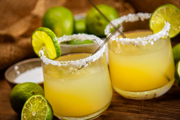 This is a photograph of two modern margarita glasses with a rim of salt surrounded by fresh cut limes on a retro wood background