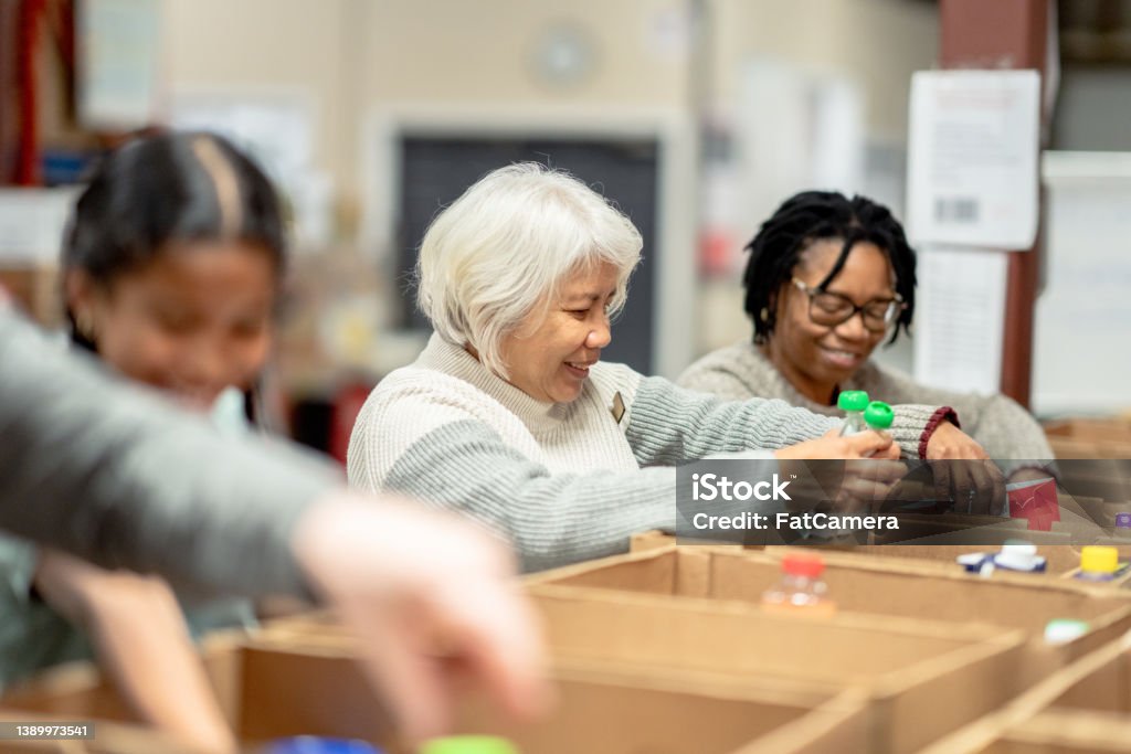Volunteers Packing at a Local Food Bank A small group of four volunteers are seen standing at a table full of cardboard boxes as they pack them with non-perishable food items at a local Food Bank.  A young mixed race girl is working with three seniors as they happily give of their time for  the cause and carefully pack the items into the boxes. Food Bank Stock Photo