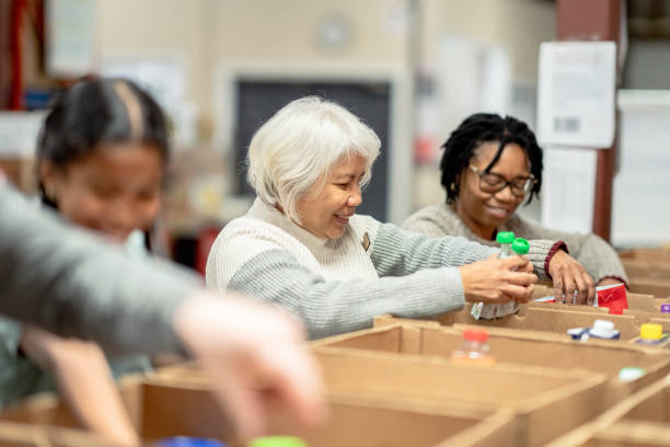 Volunteers Packing at a Local Food Bank A small group of four volunteers are seen standing at a table full of cardboard boxes as they pack them with non-perishable food items at a local Food Bank. A young mixed race girl is working with three seniors as they happily give of their time for the cause and carefully pack the items into the boxes. Senior Volunteering: Making a Difference at Any Age stock pictures, royalty-free photos & images