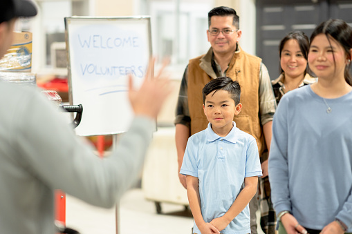 A small family of four, consisting of a mother and father and two children, is seen waiving as they enter a local food bank to volunteer their time to help packing items. They are each dressed casually and are smiling as they waive at the coordinator greeting them, and happily give of their time for the cause.