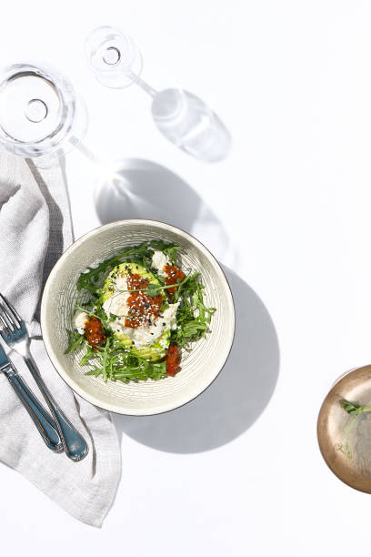 Avocado and stracciatella cheese salad. Fancy dinning with Italian salad bowl on white table with simple contemporary decor. Sunlight and harsh shadow still life. Avocado, cheese and tomato salad.