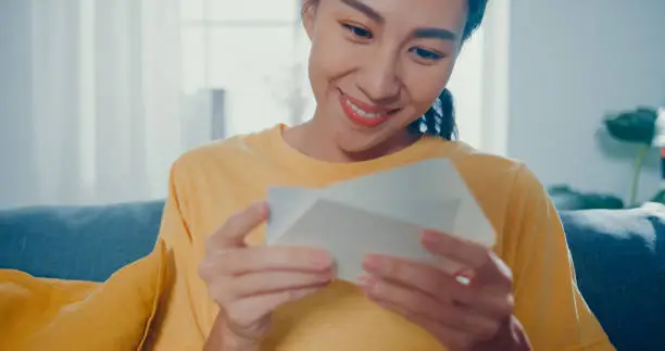 Photo of Young Asian woman feel excited unpacking gift box sitting on couch in living room at home.