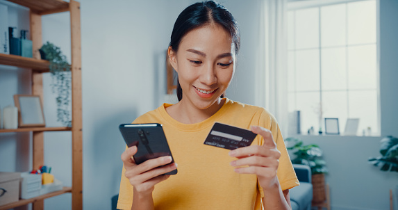 Young Asian woman holding credit card and using phone making payment online at desk in living room at home.