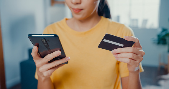 Close up of Young Asian woman holding credit card and using phone making payment online at desk in living room at home.