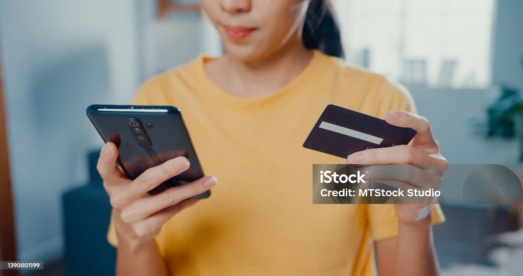 Close up of Young Asian woman holding credit card and using phone making payment online sitting at desk in living room at home. Close up of Young Asian woman holding credit card and using phone making payment online at desk in living room at home. Credit Card Stock Photo