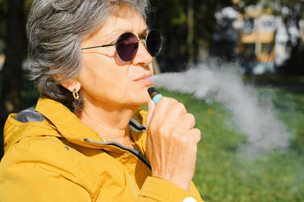 Profile portrait of elderly woman smoking modern new electronic cigarette vape, nicotine substitute, senior woman exhaling flavored vapor in park outdoors. Bad habit, pensioner and tobacco device stock photo