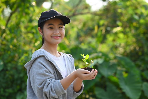 Cute girl holding young plant while standing against green nature background. Saving the world, earth day concept.