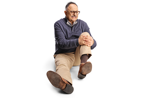 Mature man holding his painful knee and sitting on the floor isolated on white background