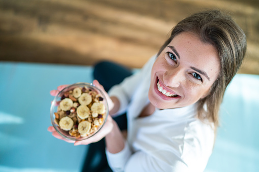 High angle view of sporty young woman holding a bowl of muesli and banana and looking at camera