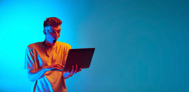 istock Studio shot of young emotional man, student using laptop isolated over blue studio background. Action, sport, fashion 1390040150
