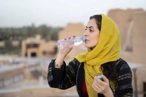 Waist-up view of mid 30s woman in traditional Middle Eastern attire standing at viewpoint and taking a drink of water to keep hydrated. At-Turaif ruins in background. Property release attached.