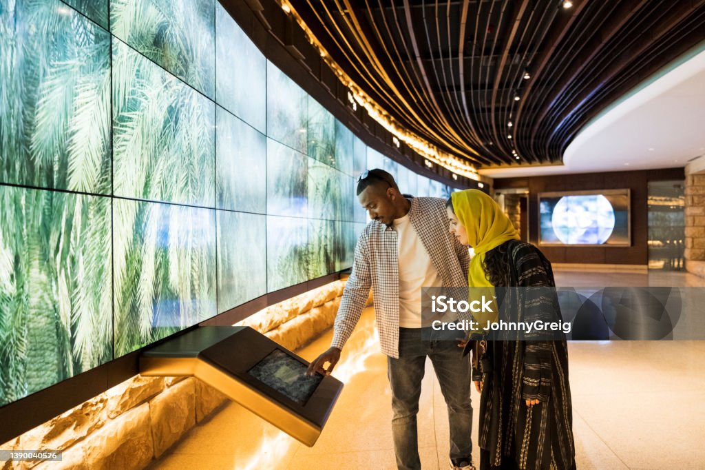 Tourists using technology in At-Turaif visitor’s centre Side view of mid adult couple enjoying interactive learning experience in UNESCO World Heritage Site near Riyadh. Property release attached. Museum Stock Photo