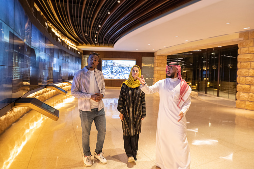 Front view of Middle Eastern man in traditional attire walking through At-Turaif visitor’s centre with couple in their 30s and discussing national heritage. Property release attached.