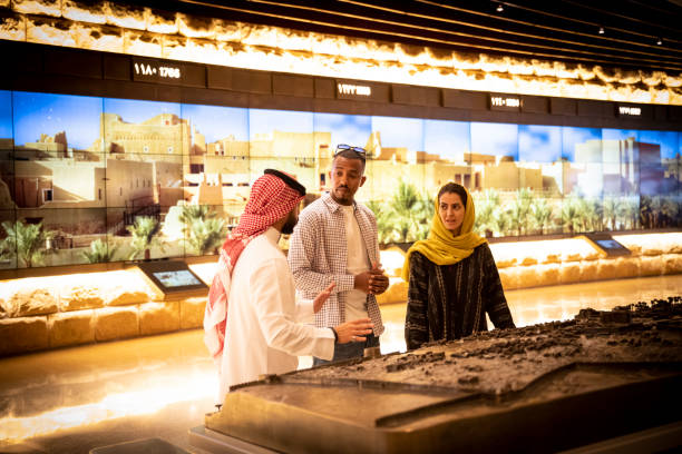 Local guide educating tourists in At-Turaif visitor’s centre Waist-up view of Middle Eastern man in traditional attire describing history of first Saudi capital and original home of royal family to mid adult couple. Property release attached. historical museum stock pictures, royalty-free photos & images