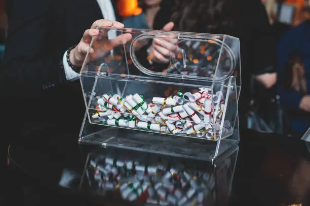 Photo of Process of prize drawings, extracting a winning numbers of lottery machine, raffle drum with bingo balls and winning tickets on event with a host and hands on lottery machine