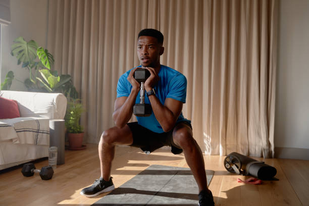 hombre joven afroamericano haciendo ejercicio desde casa concentrándose mientras está en cuclillas con una mancuerna en la sala de estar, entrenando en casa - ewan fotografías e imágenes de stock