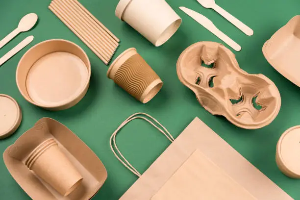 Photo of Kraft paper utensils on green background. Paper cups and containers, wooden cutlery. Street food paper packaging, recyclable paperware, zero waste packaging concept. Mockup, flat lay