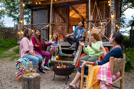 Group of senior women staying at a log cabin in the North East of England enjoying a staycation together. They are sitting outside around a fire pit talking and laughing together in summer.