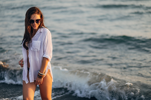 A carefree millennial woman in an unbuttoned white shirt and sunglasses enters the sea to swim