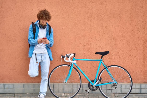 istock Young curly-haired student with headphones standing on wall outdoors using cell phone next to her bicycle 1390062146