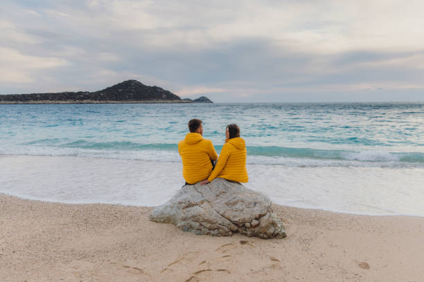 Enjoying the best time together Young happy couple of woman and man in yellow jackets sitting on the rock at the beautiful beach with island view during sunset in Turkey man walking a pug dog stock pictures, royalty-free photos & images