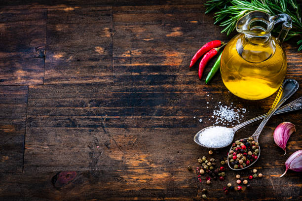 Cooking and seasoning background: high angle view of a bottle with extra virgin olive oil and two spoons with peppercorns and salt arranged at the right of a rustic wooden table leaving useful copy space for text and/or logo at the left. High resolution 42Mp studio digital capture taken with Sony A7rII and Sony FE 90mm f2.8 macro G OSS lens