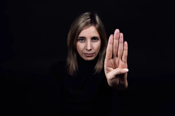 Young woman asking for help from domestic assault and abuse by showing stop sign with hand looking at camera on black background. Relative aggression. Gaslighting.