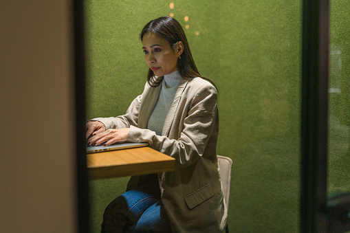 Asian businesswoman in early 40s using a small guest office in a busy corporate building. She is using a laptop while preparing for a business meeting.