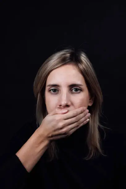 Depressed blonde woman in black blouse with hand covering mouth and making her be silent on black background. Victim of physical and psychological abuse. Gaslighting. Relative aggression.