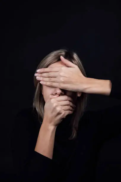 Oppressed woman with hands hiding eyes and mouth wearing black blouse on black background. Victim of physical and psychological abuse. Gaslighting. Relative aggression. Copy space.