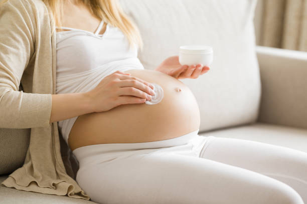 junge erwachsene schwangere frau, die zu hause auf dem sofa sitzt. hände halten glas und tragen weiße feuchtigkeitscreme auf den großen bauch auf. kümmern sie sich um perfekte, weiche und glatte körperhaut in der schwangerschaftszeit. seitenansicht. - human pregnancy abdomen naked human hand stock-fotos und bilder