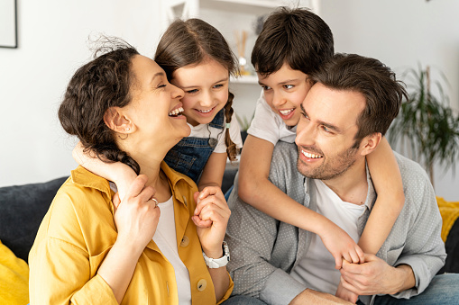 Happy together. Portrait of cheerful multiracial family of four - mom, dad, son and toddler daughter sitting on the sofa in living room, looking to each other and laughing, enjoying weekend at home