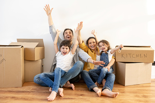 Overjoyed multiracial family of four sitting on floor surrounded cardboard boxes in empty living room, cute daughter holding keys with keychain in form of little house. Owners of estate, mortgage