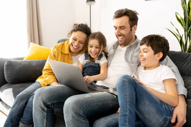 alegre familia feliz de cuatro usando computadora portátil sentada en el sofá en acogedor apartamento - child computer internet laptop fotografías e imágenes de stock