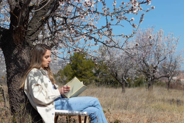 istock Young woman leaning on tree trunk with flowers reading a book relaxed 1390081217