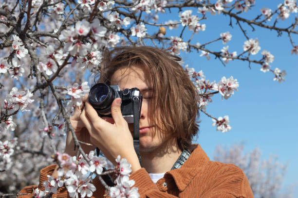 Portrait of young long haired man taking pictures with a film camera flowers Close up portrait of young long haired man taking pictures with a film camera to beautiful cherry blossom flowers under blue sky photographic film camera stock pictures, royalty-free photos & images