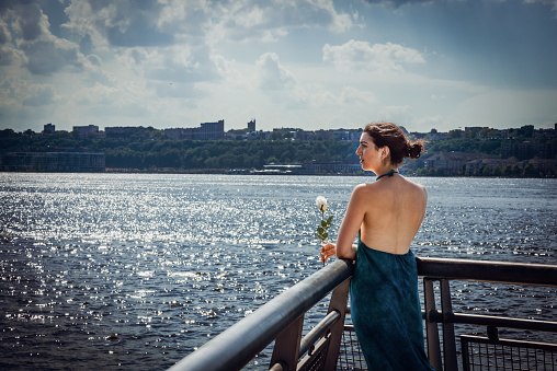 Waiting for you. Young girl with bare back, long blue dress, holding white rose, standing by river in New York City, waiting for you. \