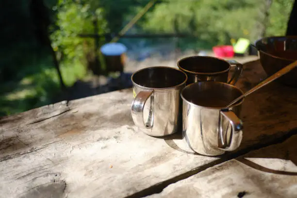 Photo of Three tourist iron mugs on the wooden table in a forest camping. Survival equipment for hiking concept. Close up photo of the 3 metal cup illuminated by the sun on timber desk. Journey, Trekking Theme