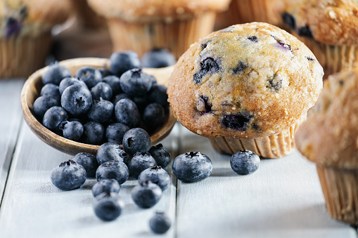 Fresh blueberry muffins with raw blueberries spilling from a wooden spoon. Selective focus on center with blurred foreground and background.