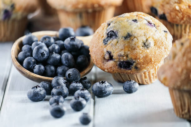 Homemade Blueberry Muffins with Ingredients Fresh blueberry muffins with raw blueberries spilling from a wooden spoon. Selective focus on center with blurred foreground and background. Blueberry Muffin stock pictures, royalty-free photos & images