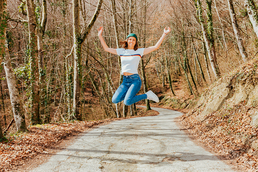 Portrait of young cheerful woman in mid air at countryside road. Happy smiling female wears green hat and lgbtiq shirt, jumps near the forest.