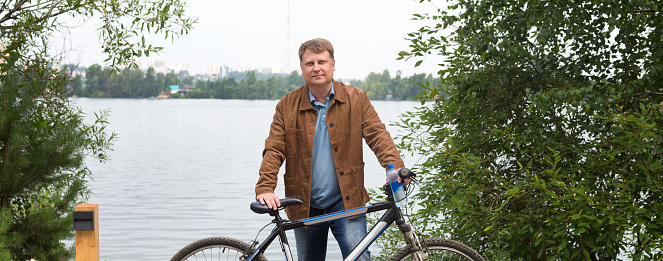 An adult blond man brown corduroy jacket is standing by the river in the park with a bicycle.