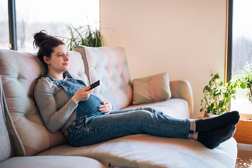 Pregnant woman on sofa at home watching TV.