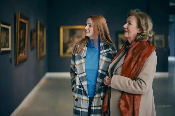 Photo of Grandmother and adolescent granddaughter are looking at the paintings in the art gallery.