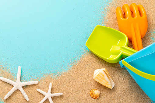 Top view of a bucket of sand, a rake and a shovel laying down on a scattered sand and surrounded by a starfish and some seashells. The sand and all objects are to the right and bottom of the image, leaving useful copy space in the top left corner against a light blue background.