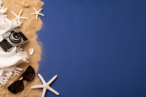 Top view of a camera laying on a beach sand surrounded by sunglasses, a beach towel, some star fish and seashells. All the objects are at the left side of the image leaving a useful copy space at the right side on a dark blue background