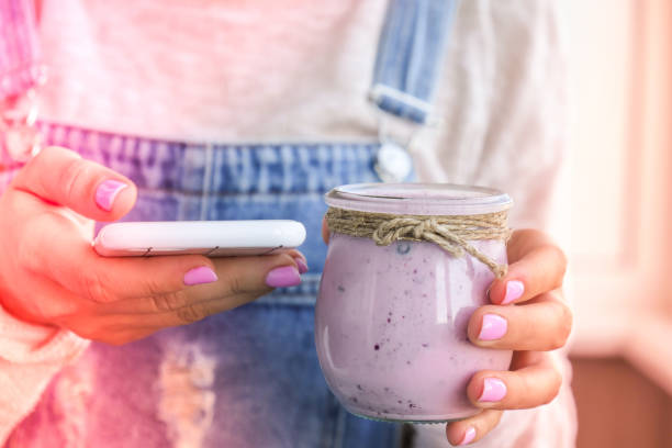 female hands holding blueberry smoothie topped with blueberries. woman drinking glass of breakfast protein smoothie drink. using mobile phone, social media, snack break, online studying, recipe - blueberry smoothie milk shake drink imagens e fotografias de stock