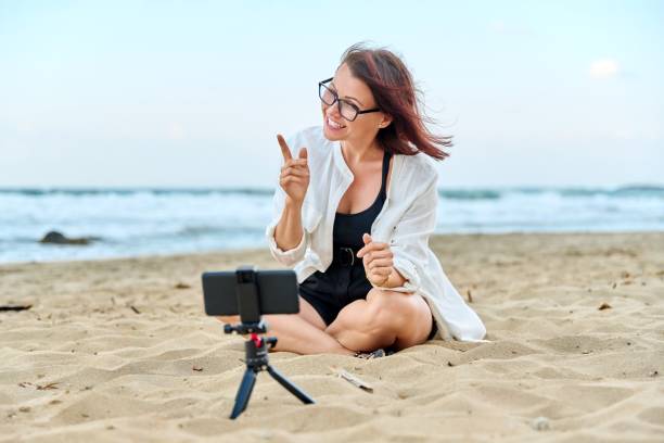 istock Middle aged woman sitting on beach with smartphone using video call 1390116771