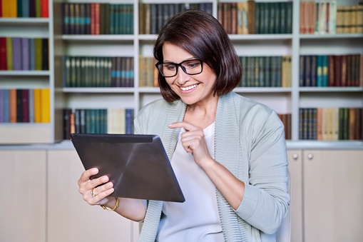 Business smiling mature woman using digital tablet in library office. Profession, occupation, technology, career, job, education, people concept