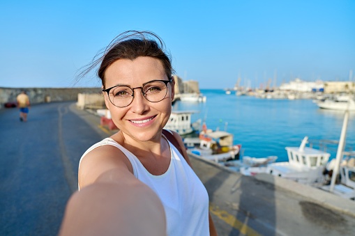 Close up face of happy middle aged woman taking selfie portrait. Smiling female with backpack enjoying summer vacation travel trip in european maritime destination